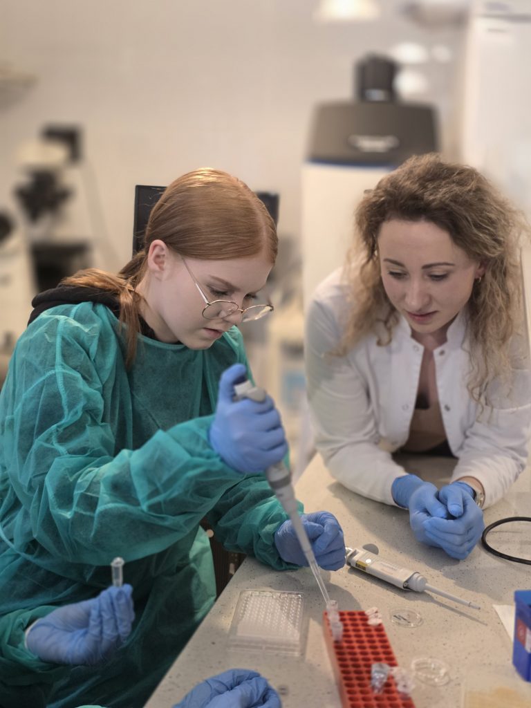 Studentka w zielonym fartuchu wykonuje pipetowanie, a prowadząca w białym kitlu uważnie obserwuje i instruuje przy stanowisku laboratoryjnym.