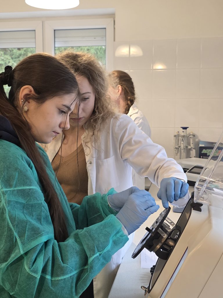 Prowadząca w białym kitlu pomaga studentce w zielonym fartuchu podczas obsługi urządzenia laboratoryjnego.