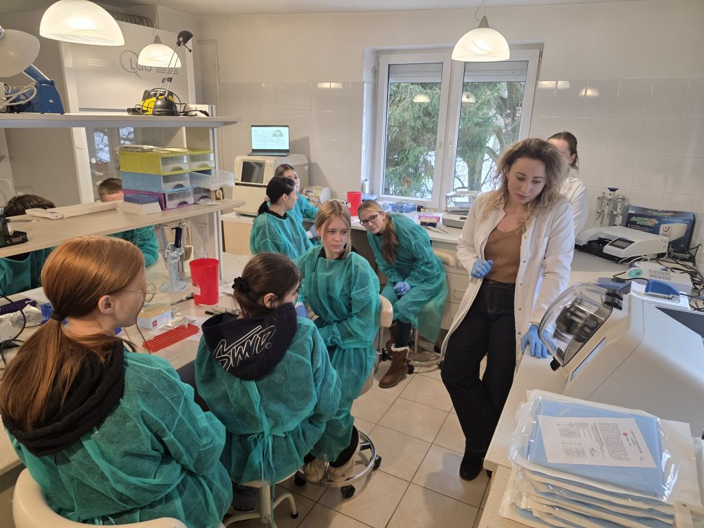 Prowadząca w białym kitlu omawia przebieg zajęć, a studenci w zielonych fartuchach siedzą przy stanowiskach w laboratorium.