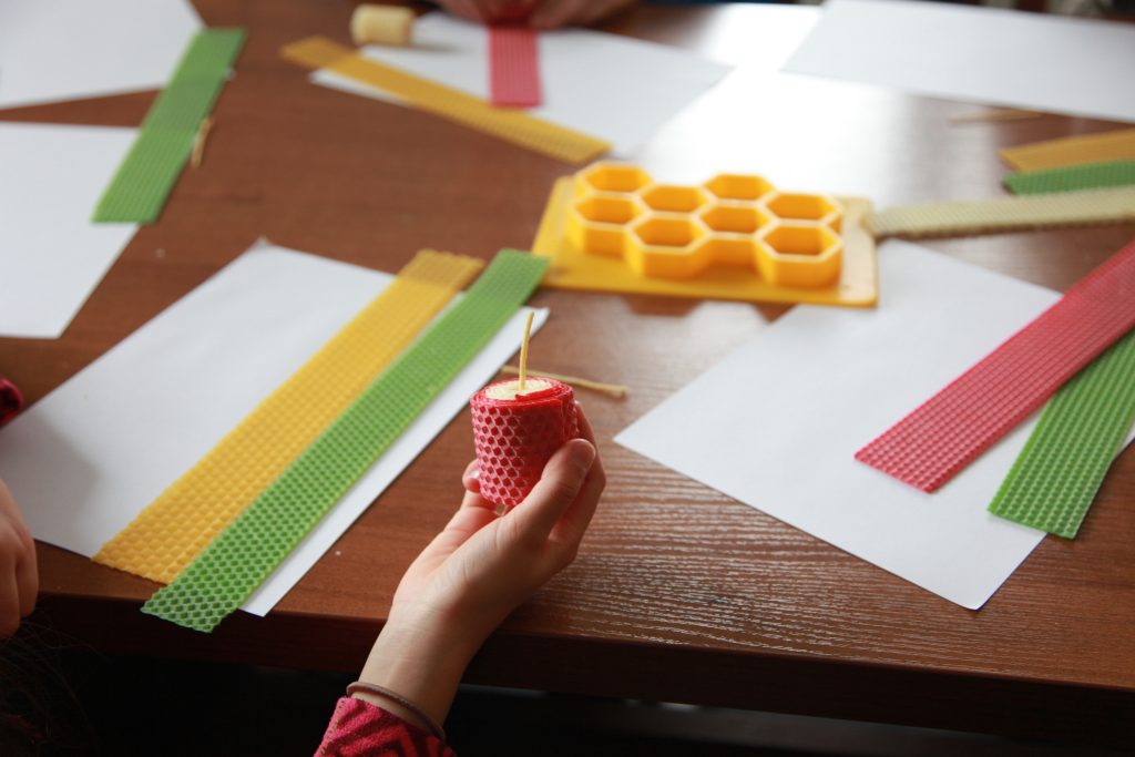 Dziecko trzyma małą świecę z wosku pszczelego, a na stole leżą kolorowe arkusze węzy i materiały do zajęć plastycznych.