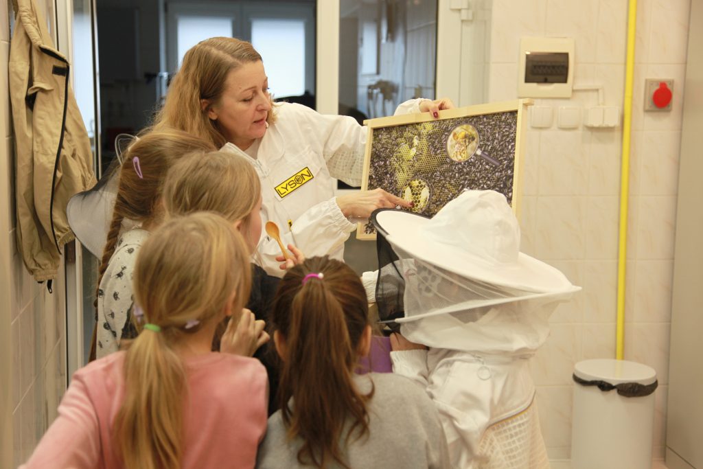 Kobieta w stroju ochronnym pokazuje dzieciom planszę z plastrem miodu i pszczołami, omawiając budowę ula podczas zajęć edukacyjnych.
