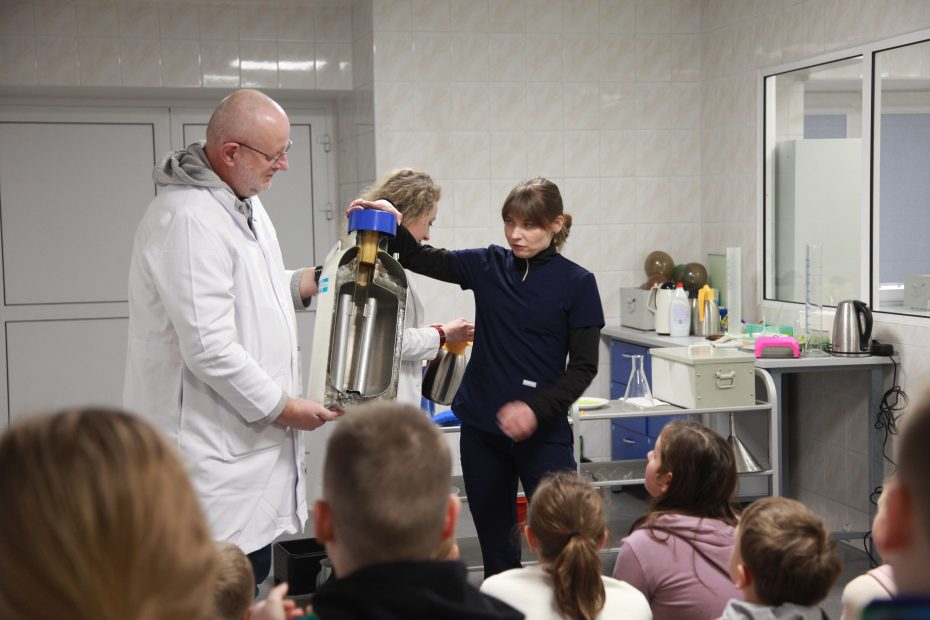 Grupa dzieci siedzi w laboratorium i z zainteresowaniem obserwuje, jak dwoje pracowników prezentuje przekrojony model urządzenia badawczego.