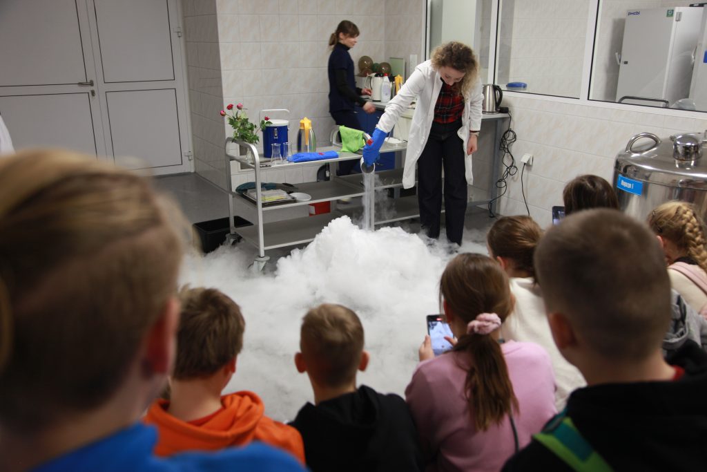 Pracownica laboratorium w fartuchu przeprowadza pokaz z użyciem ciekłego azotu, a grupa dzieci obserwuje unoszącą się nad podłogą białą mgłę.