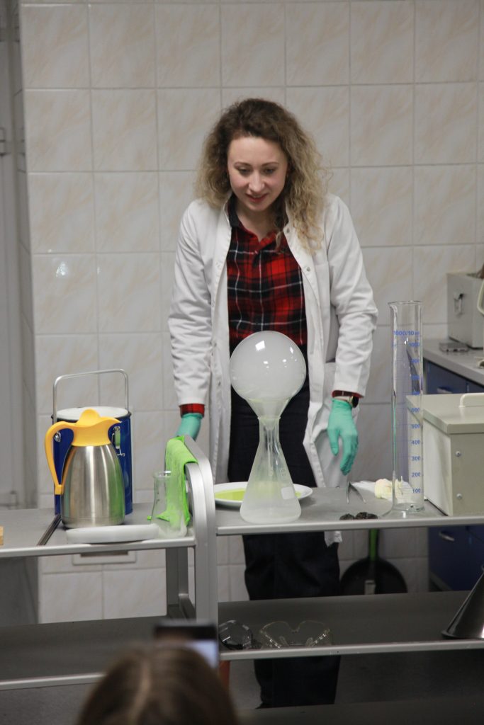 Pracownica laboratorium w fartuchu i rękawicach prezentuje doświadczenie chemiczne, obserwując unoszącą się nad kolbą bańkę z pary.