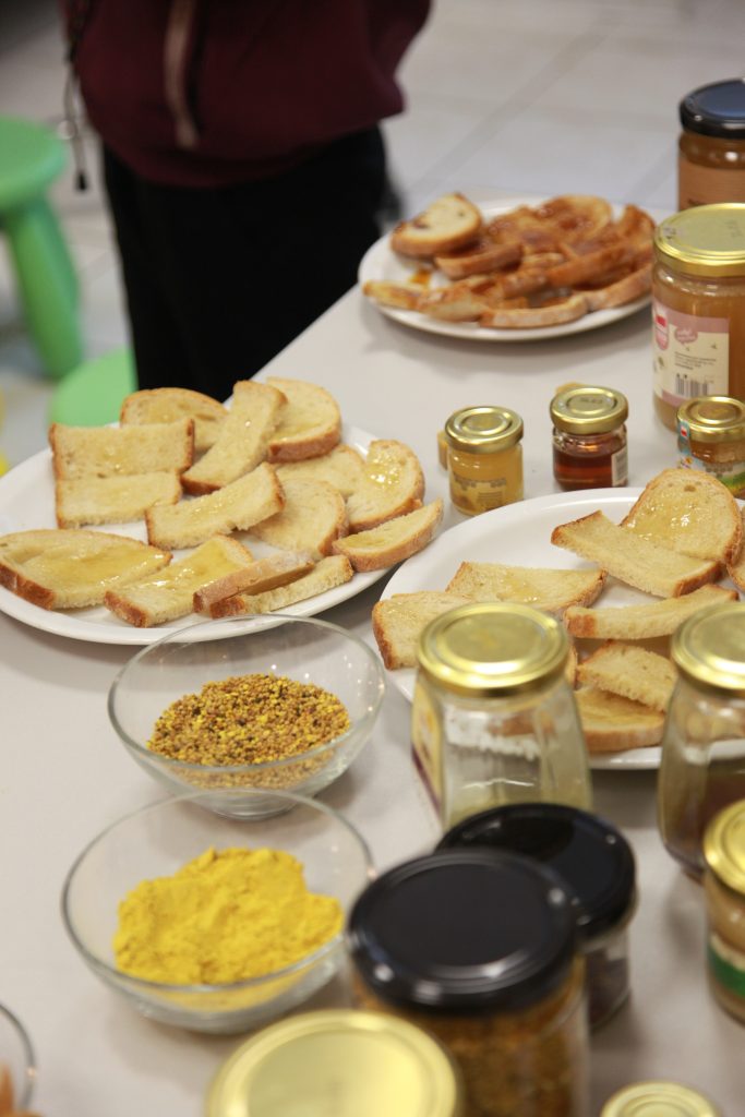 Na stole ustawiono talerze z kawałkami pieczywa posmarowanymi miodem, obok słoiki z różnymi jego rodzajami oraz miseczki z pyłkiem pszczelim i innymi produktami pszczelimi do degustacji.