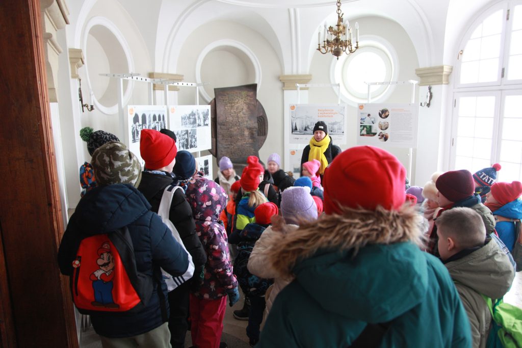 Grupa dzieci w zimowych czapkach i kurtkach stoi w jasnej sali wystawowej, słuchając przewodnika na tle plansz z fotografiami i opisami.