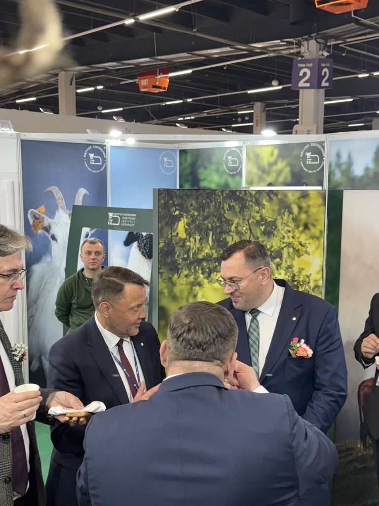 Grupa mężczyzn w garniturach rozmawia przy stoisku targowym związanym z rolnictwem. W tle materiały promocyjne i grafiki zwierząt gospodarskich.