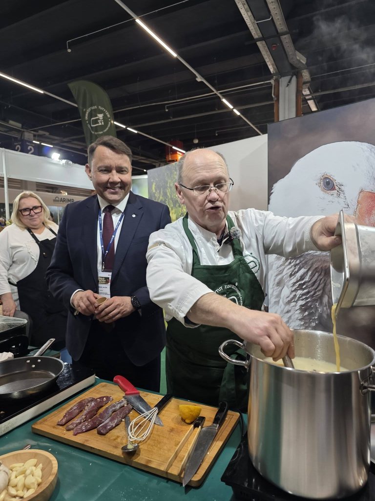 Mężczyzna w fartuchu kucharskim przygotowuje potrawę na stoisku targowym, w tle obserwują go uczestnicy wydarzenia.