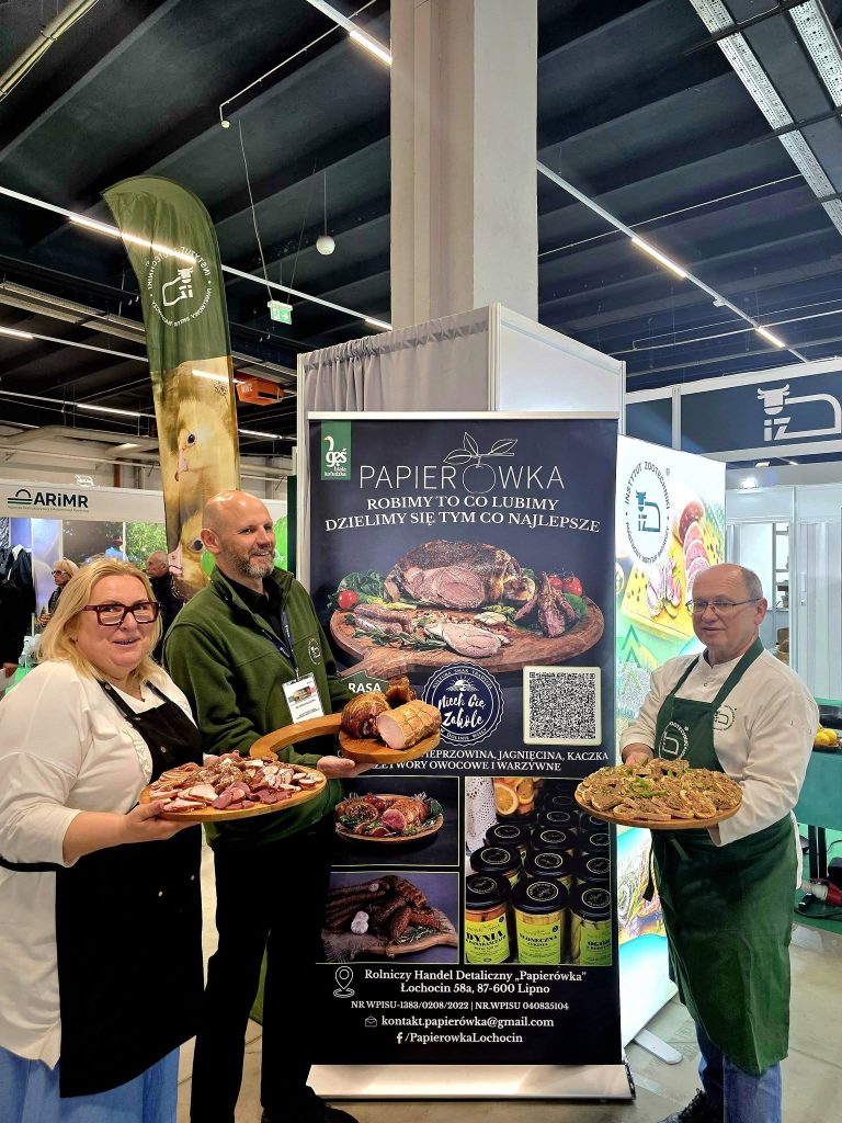 Trzy osoby prezentują półmiski z wędlinami i potrawami regionalnymi na stoisku targowym z materiałami promocyjnymi w tle.