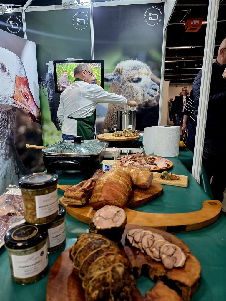 Stoisko targowe z regionalnymi wyrobami mięsnymi; na pierwszym planie deski z wędlinami, w tle osoba przygotowująca potrawy.