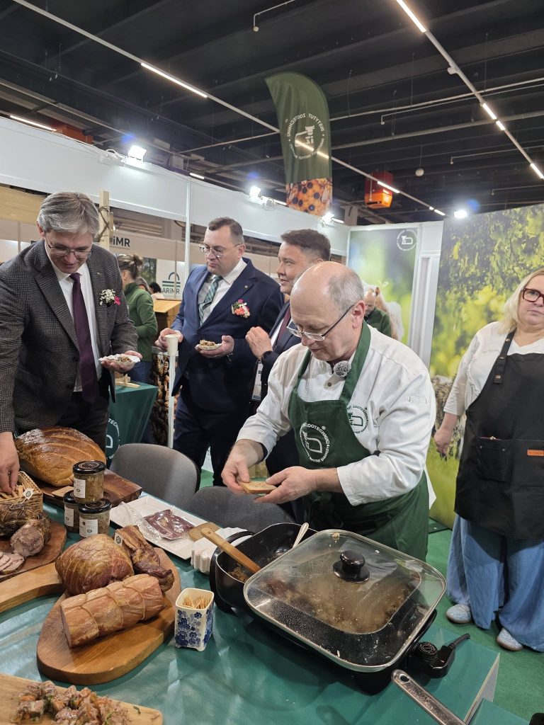 Degustacja produktów regionalnych na stoisku targowym; kucharz przygotowuje kanapki, a uczestnicy próbują potraw.