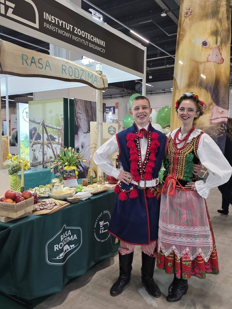 Dwoje młodych ludzi w strojach ludowych stoi przy stoisku „Rasa Rodzima” Instytutu Zootechniki, obok ekspozycji regionalnych produktów.