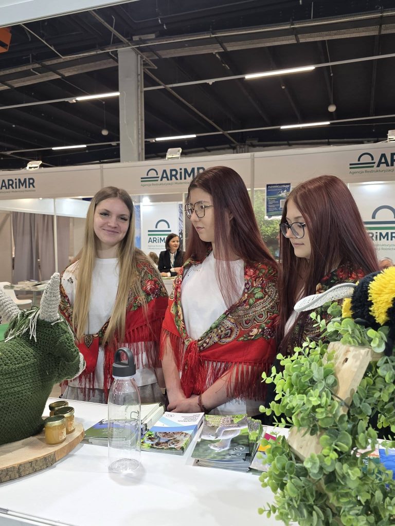 Trzy kobiety w strojach inspirowanych folklorem prezentują materiały informacyjne i produkty na stoisku targowym.